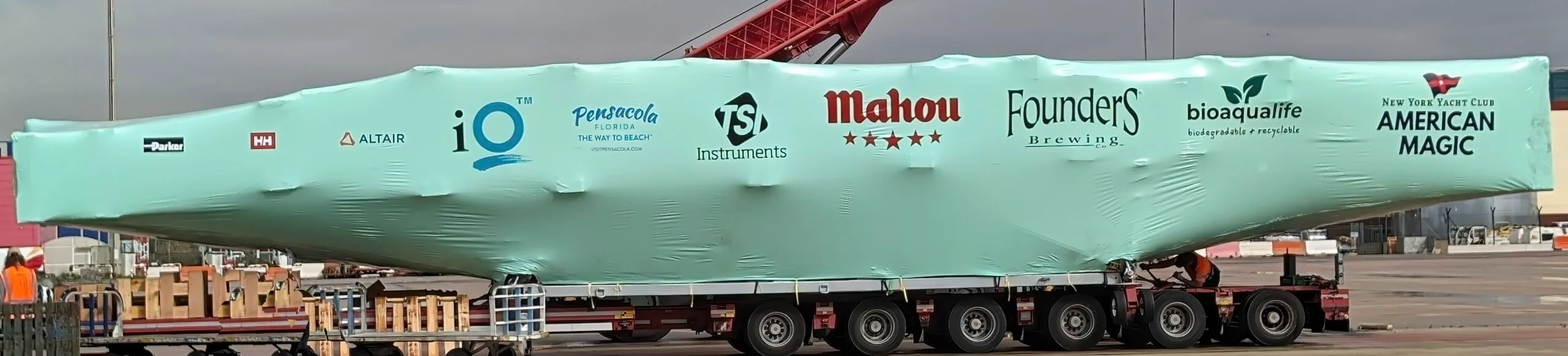 bioaqualife: A large boat covered in a light green protective wrap is being transported on a flatbed truck. The wrap displays various sponsor logos, including Altair, Mahou, Founders Brewing, and American Magic. The background shows an industrial area with cloudy skies.