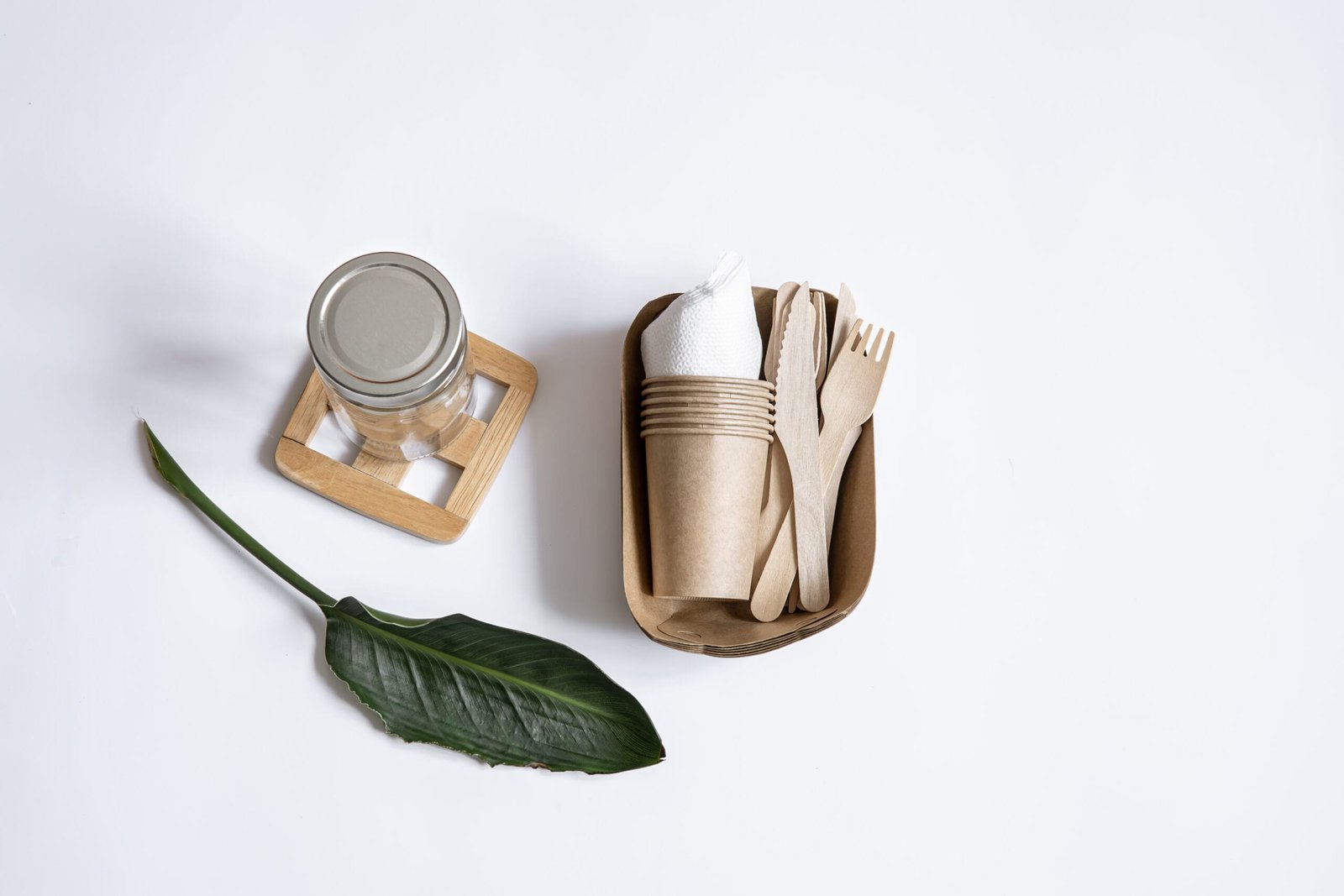 bioaqualife: Eco-friendly dining set featuring a reusable glass jar with a lid on a wooden coaster, a brown rectangular tray with compostable cups, a fork, and a knife on a white background. A green leaf adorns the set, enhancing the natural theme.