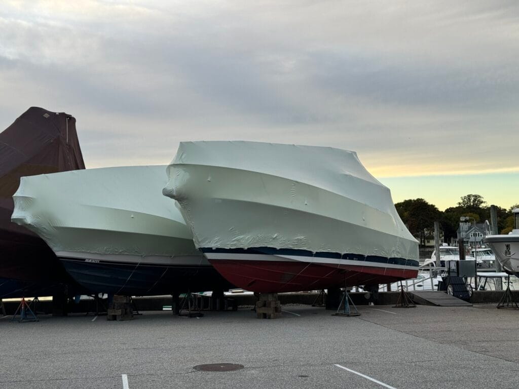 bioaqualife: Several boats covered in white protective wrapping are parked on supports in a marina. The sky is overcast, and trees are visible in the background.