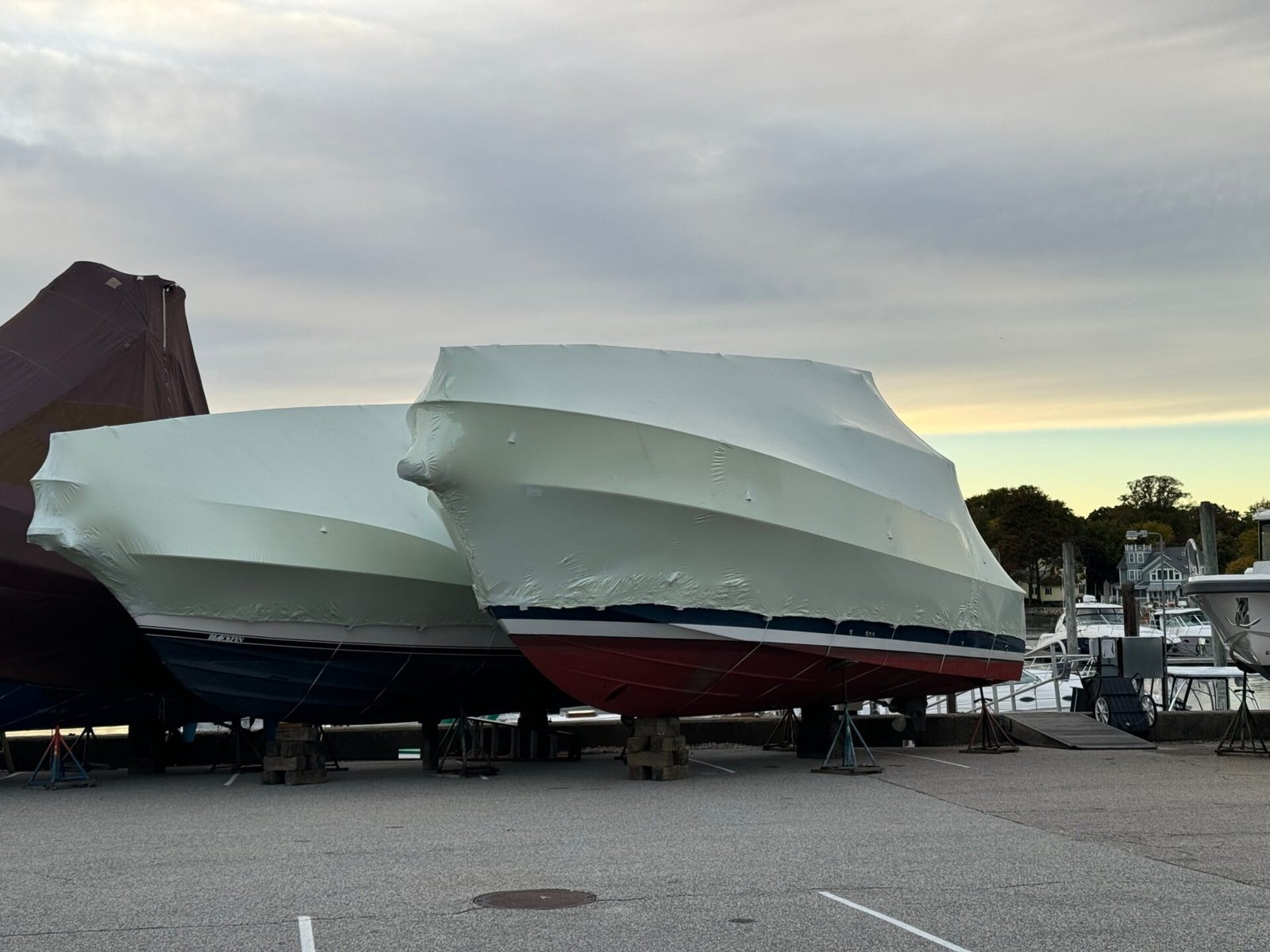 bioaqualife: Several boats covered in white protective wrapping are parked on supports in a marina. The sky is overcast, and trees are visible in the background.
