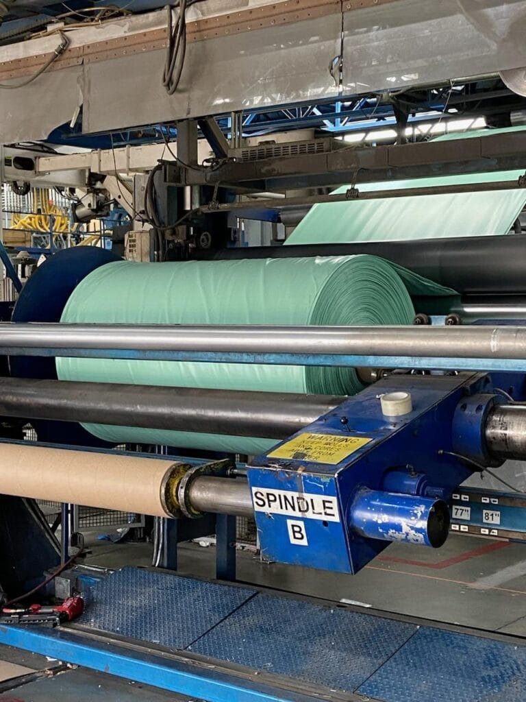 bioaqualife: Industrial machinery with large rolls of light green fabric being processed. The machine includes a blue section labeled "SPINDLE B" and is set in a factory environment with metal structures and a blue platform.