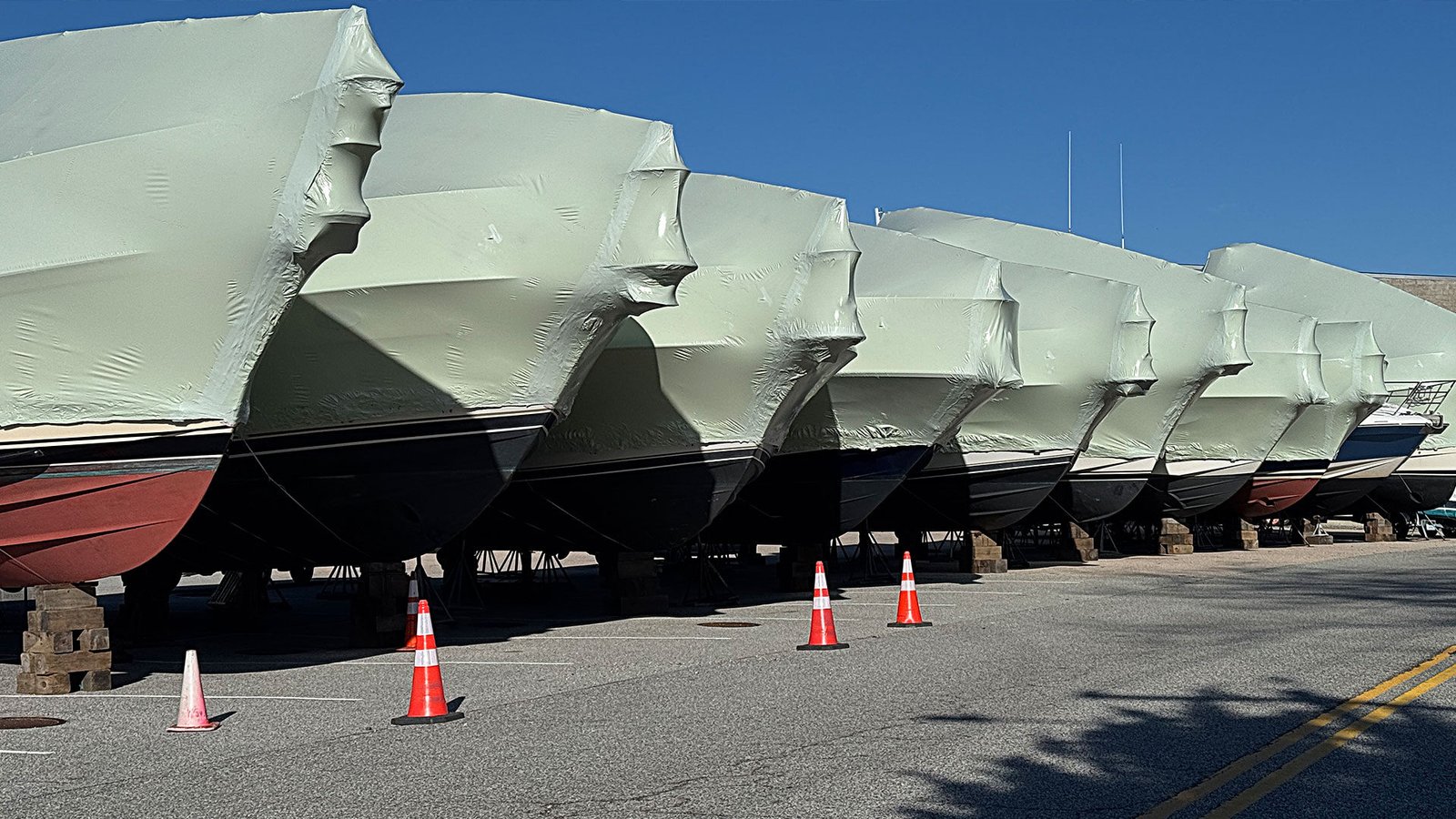 bioaqualife: Several boats covered in white protective wraps are lined up on blocks in a parking lot. Orange traffic cones are placed along the edge, and the sky is clear and blue.