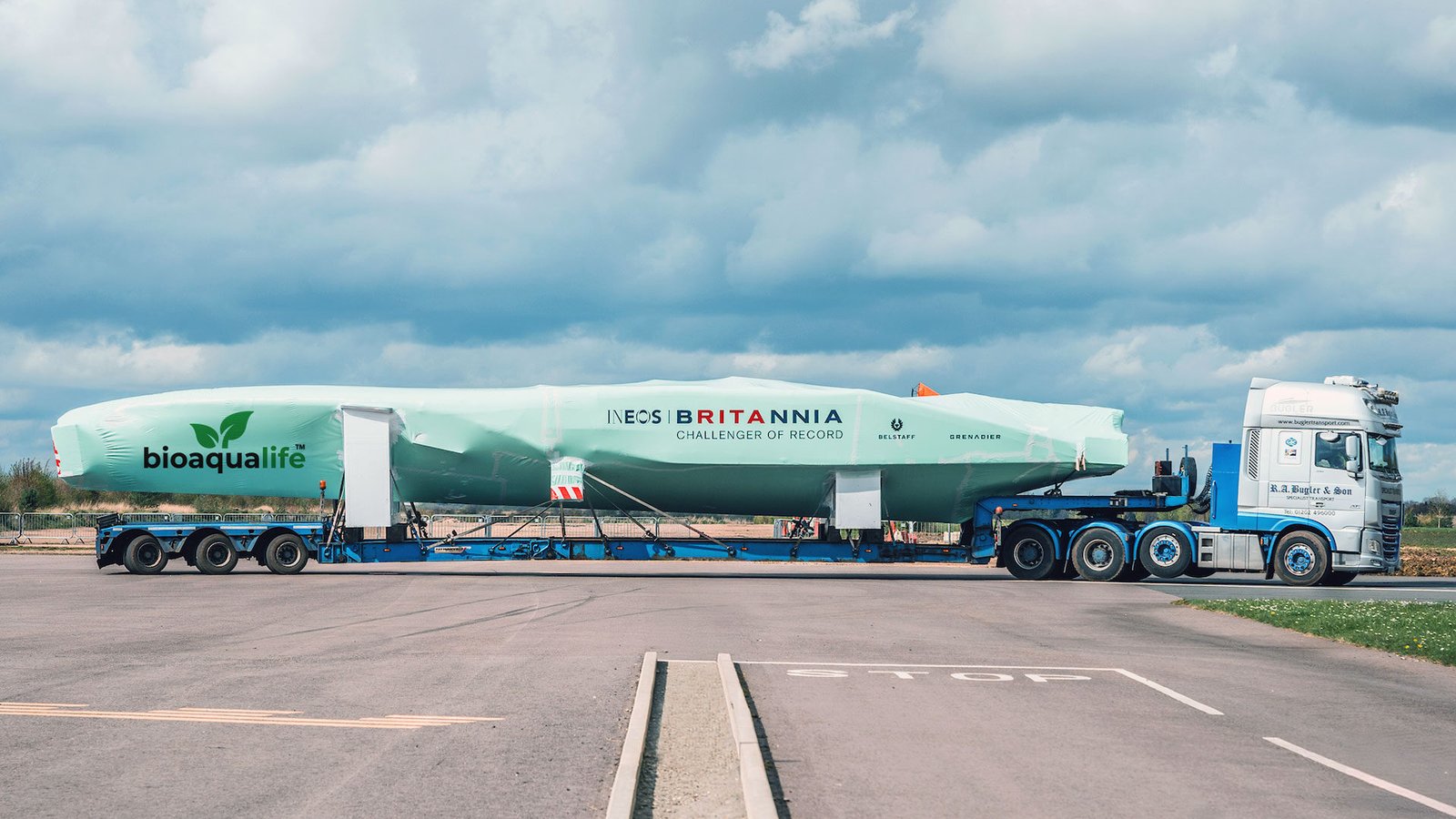 bioaqualife: A large truck transports a long, covered sailboat mast labeled "INEOS BRITANNIA" and "bioaqualife" on a flatbed trailer along a paved road under a cloudy sky.