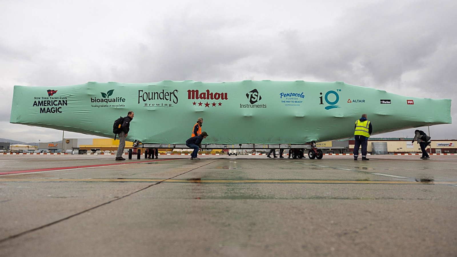 bioaqualife: People in safety vests move and inspect a large, teal, covered object on an airport tarmac. The cover displays various sponsor logos, including American Magic, Founders, Mahou, and others. The sky is overcast.