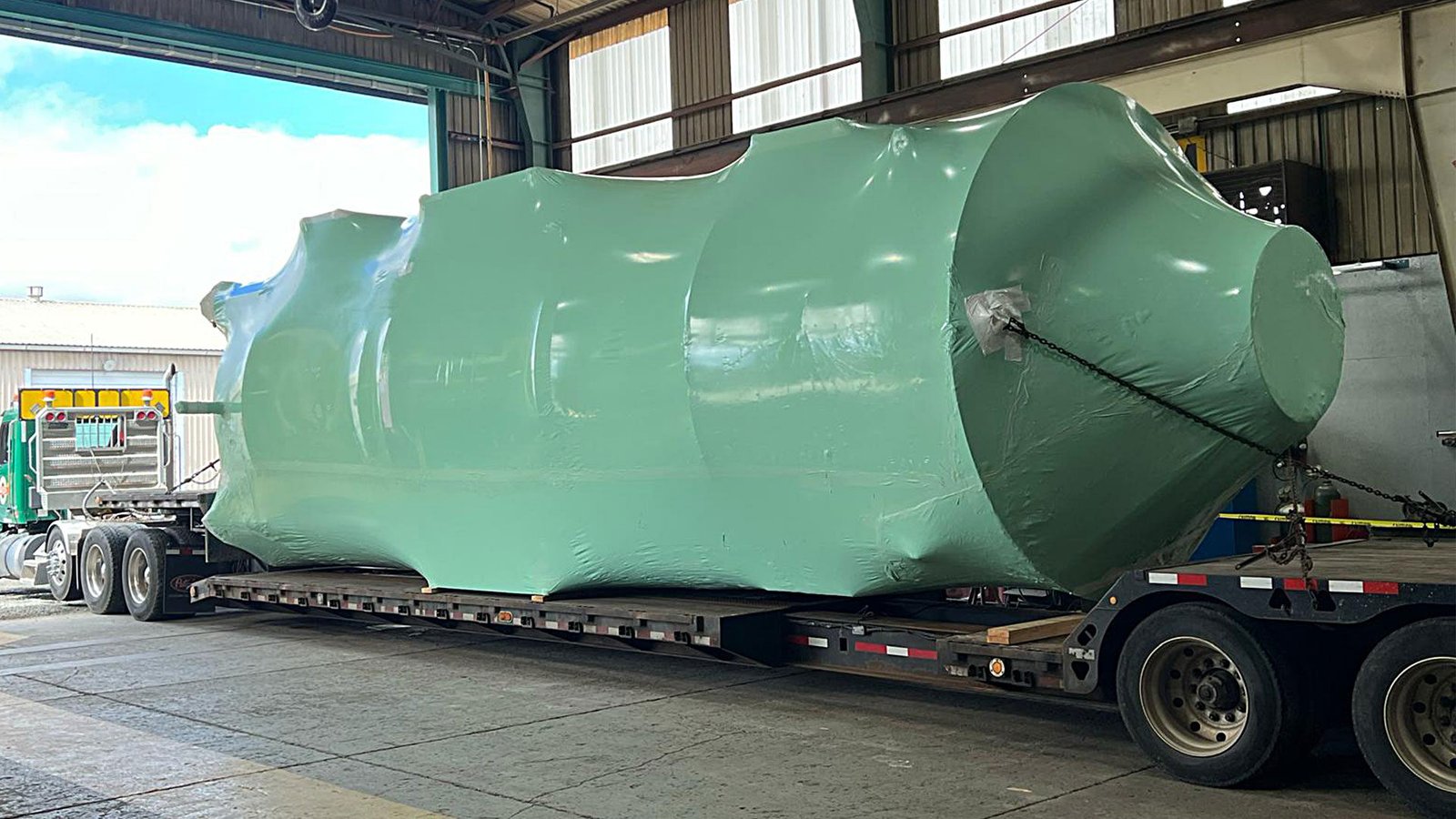 bioaqualife: A large, green-wrapped industrial tank is secured on a flatbed trailer inside a warehouse, ready for transport. Sunlight filters in through the windows, illuminating the scene.