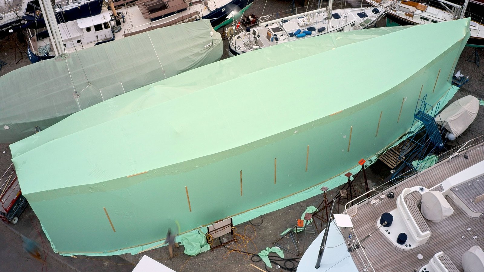 bioaqualife: A large boat is covered with green plastic sheeting at a shipyard, surrounded by other boats and equipment. The view is from above, showing the entire covered vessel and parts of the surrounding dock area.