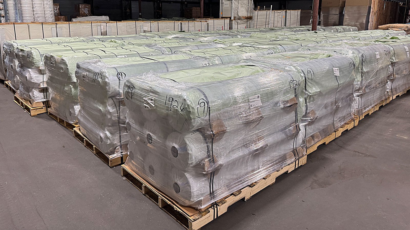 bioaqualife: Stacks of large, tightly wrapped rolls of material on wooden pallets are arranged in rows inside a warehouse with a concrete floor and industrial shelving in the background.