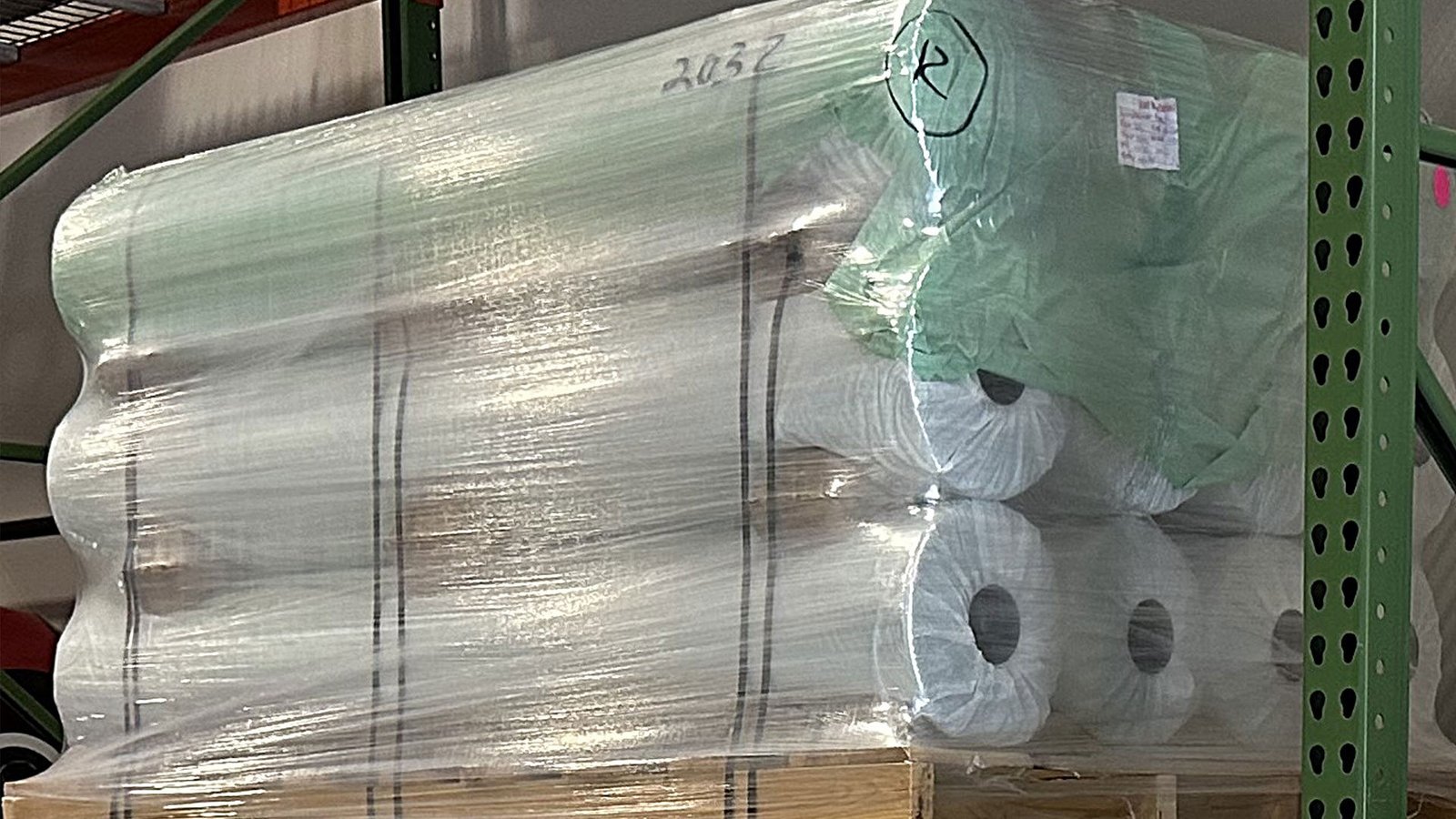 bioaqualife: A stack of large rolls of white fabric wrapped in clear plastic sits on a wooden pallet in a warehouse storage rack. Handwritten markings and labels are visible on the packaging.