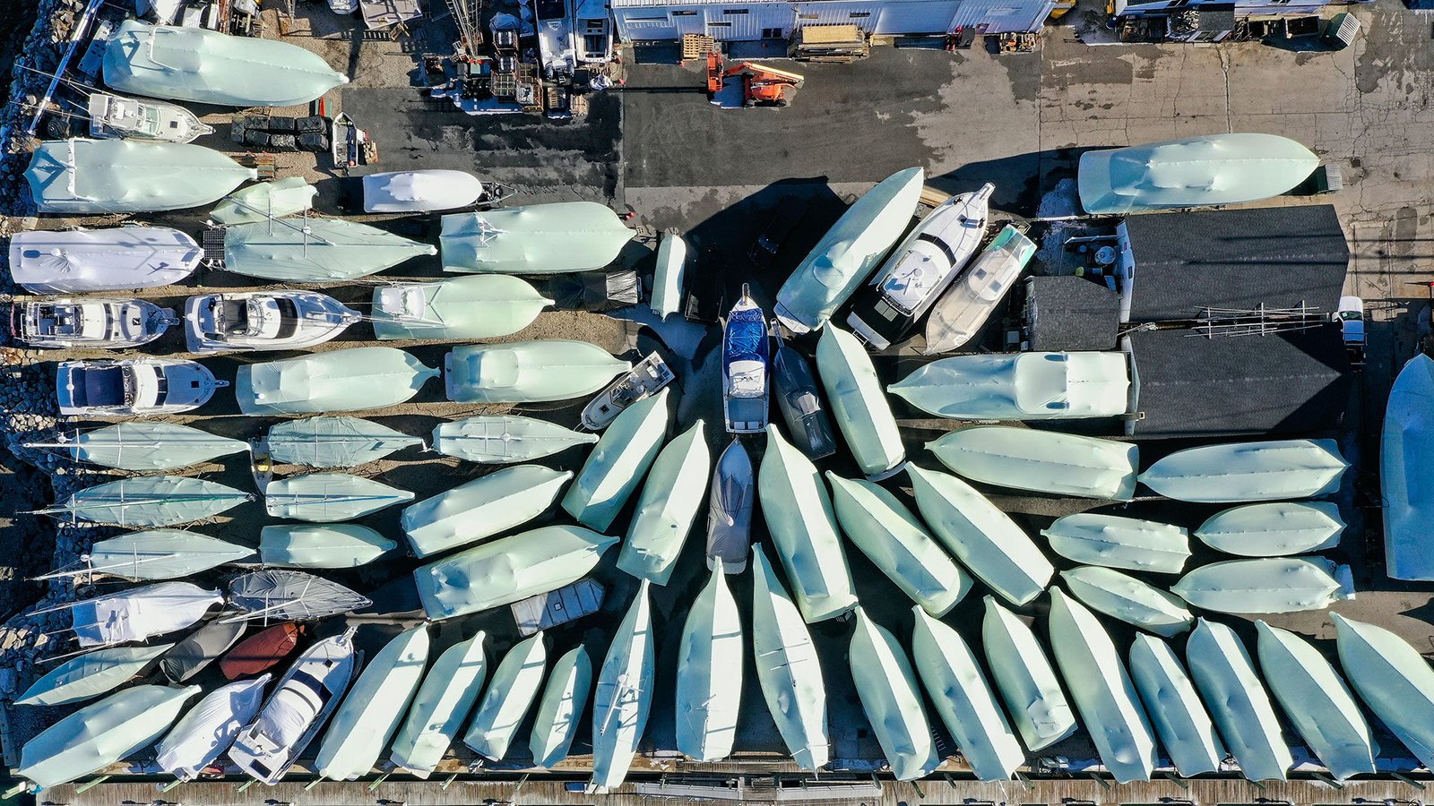 bioaqualife: Aerial view of numerous boats, mostly covered with white tarps, arranged in rows in a boatyard next to buildings and equipment on a paved lot.