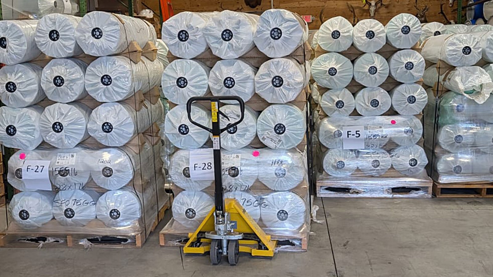 bioaqualife: Stacks of large rolls of white material wrapped in plastic are arranged on wooden pallets in a warehouse. A yellow pallet jack is positioned in front of the pallets. Each stack is labeled with tags.