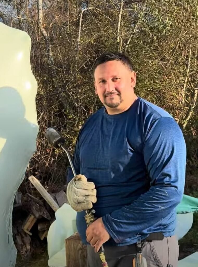 bioaqualife: A man in a blue long-sleeve shirt and gloves holds a propane torch outside, standing next to a large ice sculpture with trees and sunlight in the background.