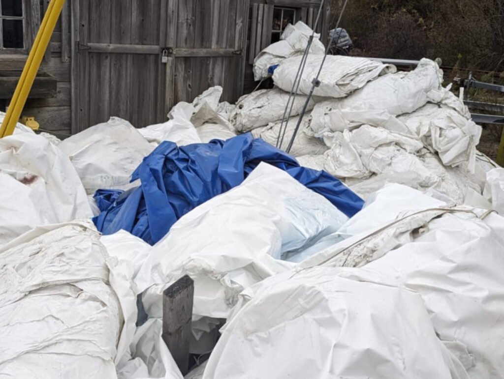 bioaqualife: A large pile of crumpled white and blue tarps or plastic sheeting lies outdoors next to a weathered wooden building. Various poles and wooden boards are scattered among the piles.