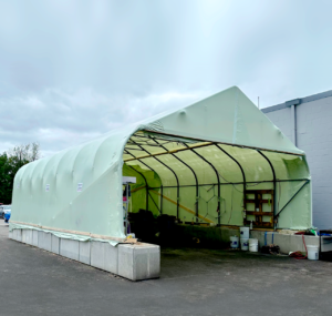 bioaqualife: A large green fabric-covered shelter with an open front stands on a concrete platform beside a gray building. Various tools, shelves, and items are visible inside the structure. The sky is overcast.