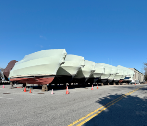 bioaqualife: Several boats covered in white shrink wrap are lined up on land in a parking lot, surrounded by orange traffic cones, under a clear blue sky.