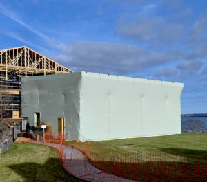 bioaqualife: A large building under construction is wrapped in white protective sheeting. Wooden framework is visible on the roof. A red safety fence surrounds the area, and there is a pathway and grass in the foreground, with water in the background.
