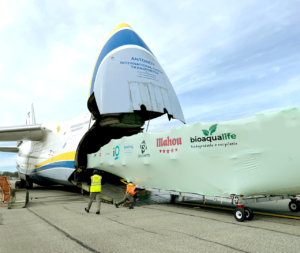 bioaqualife: Two workers guide a large, covered cargo load being unloaded from the nose of an Antonov cargo plane. The load has logos for IO, Mahou, and Bioqualife. The plane&rsquo;s nose is raised open on an airport tarmac.