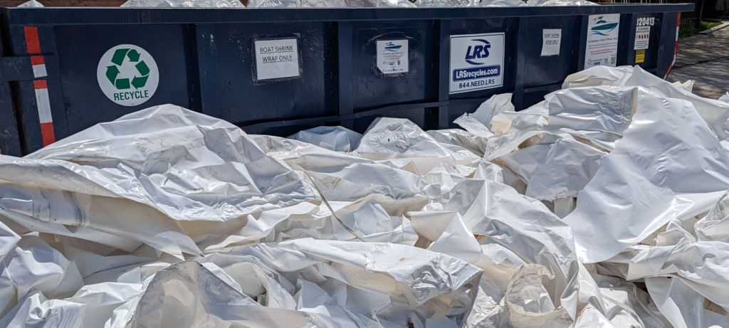 bioaqualife: A large blue recycling dumpster is filled with crumpled white plastic sheets. Several signs are posted on the dumpster, including a green recycle symbol and company information.
