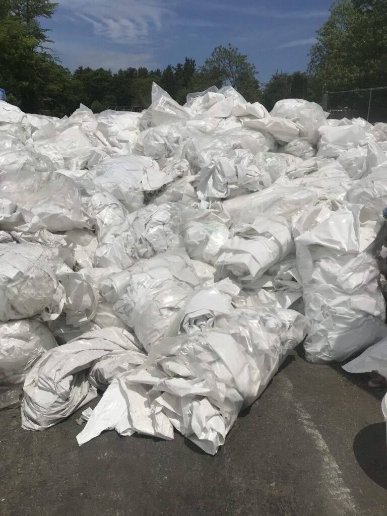 bioaqualife: A large pile of white plastic bags or sheeting stacked outdoors on a paved surface, with trees and a fence visible in the background under a partly cloudy sky.