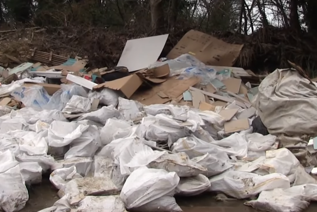bioaqualife: A large pile of white garbage bags and scattered cardboard boxes and debris is heaped outdoors near trees and bushes, indicating illegal dumping or improper waste disposal.