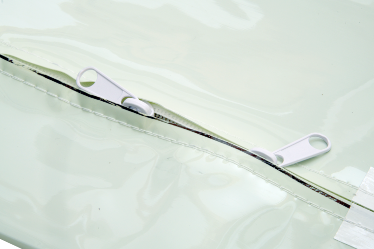 bioaqualife: A close-up of a light-colored vinyl or plastic material with a partially open zipper featuring two white zipper pulls. The stitching and shiny surface of the material are clearly visible.