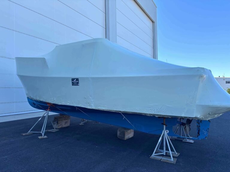 bioaqualife: A white and blue boat covered in protective shrink wrap is supported by metal stands and blocks beside a large building under a clear blue sky.