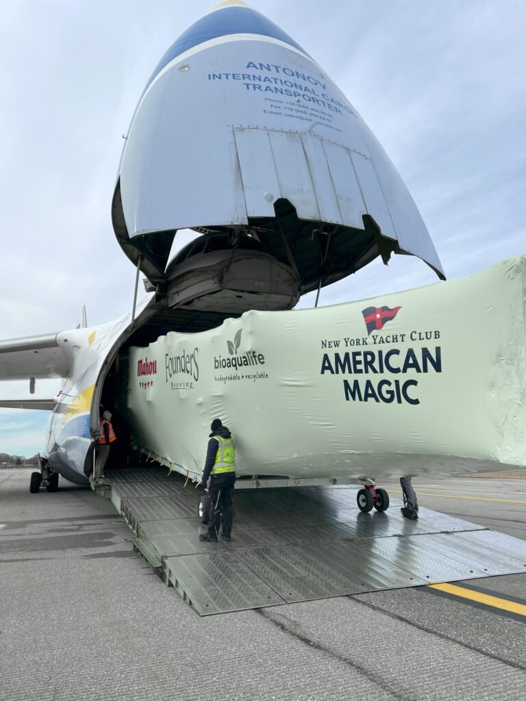bioaqualife: A large aircraft with its nose door open is unloading a covered sailboat labeled "New York Yacht Club American Magic" as workers in safety vests assist on the airport tarmac.
