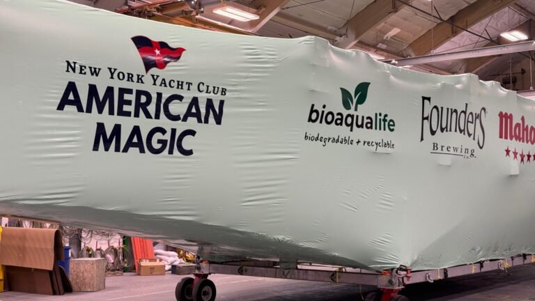 bioaqualife: A large boat covered in protective white wrap displays logos for the New York Yacht Club American Magic, bioaqualife, Founders Brewing, and Mahou inside an indoor facility.