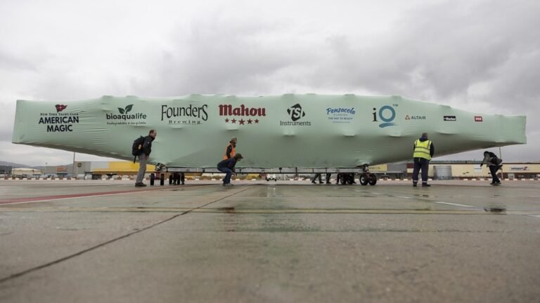 bioaqualife: Several people move a large, covered sailboat on an airport tarmac. The green cover displays logos of various sponsors including Mahou, Founders Brewing Co., and others. The sky is overcast.