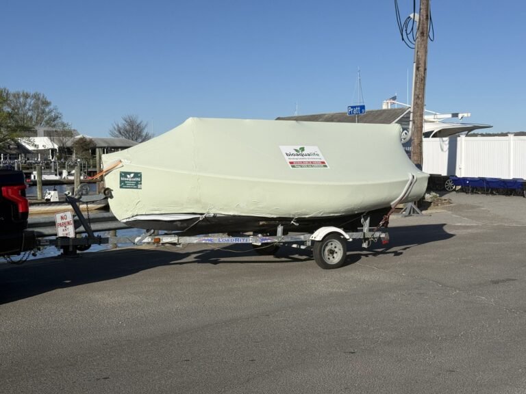 bioaqualife: A small boat covered with a light green tarp labeled "Kindervelt 16" is loaded on a trailer hitched to a vehicle, parked on an asphalt surface near a marina with docks and boats in the background.