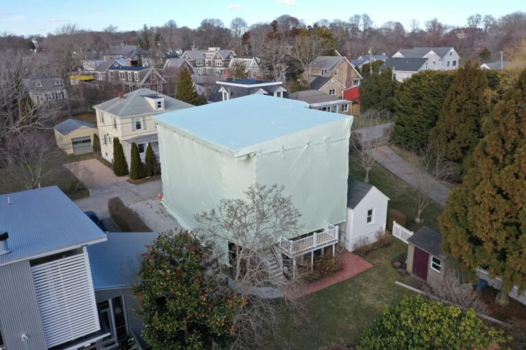 bioaqualife: A two-story house is covered by a large white tarp, fully enclosing it. The house is in a residential neighborhood with other homes, trees, and well-kept yards visible around it.