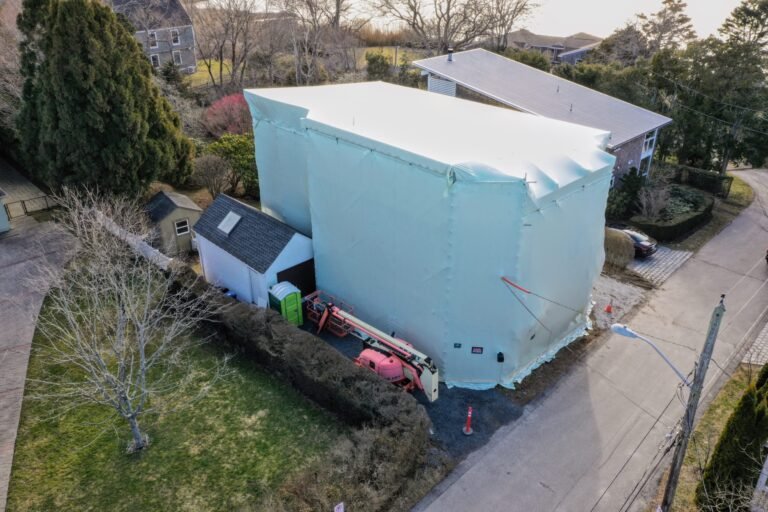bioaqualife: A large house is completely covered in white protective wrapping, likely for renovation, with a construction lift and portable toilet nearby; the house sits on a corner lot in a suburban neighborhood.