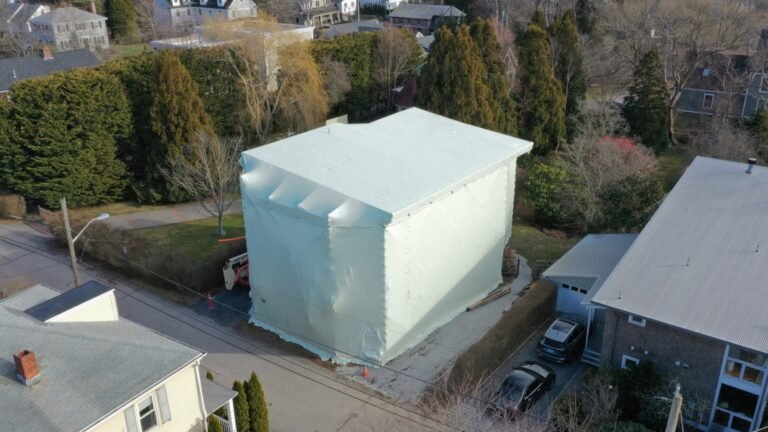 bioaqualife: A large house is fully wrapped in white plastic sheeting, covering the entire structure. The home is situated on a residential street with neighboring houses and trees surrounding it.