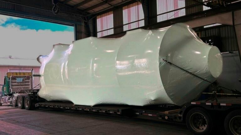 bioaqualife: A large, irregularly shaped object wrapped in white plastic is secured on a flatbed trailer inside an industrial warehouse, with sunlight streaming through windows and a green truck visible outside.
