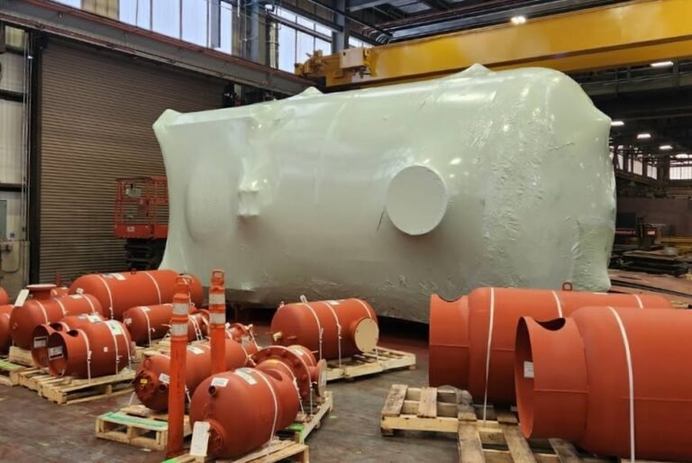 bioaqualife: Large white industrial tank wrapped in protective material stands in a warehouse. Several smaller red cylindrical tanks, secured to wooden pallets, are arranged in the foreground. The space is lit by large overhead lights.