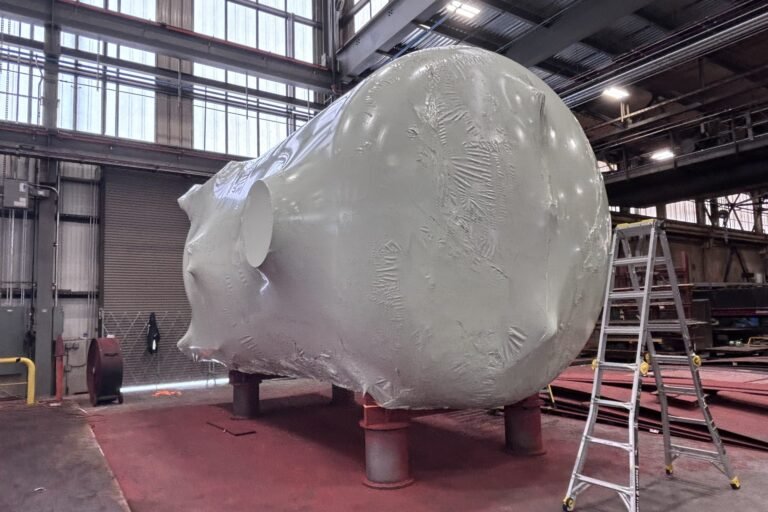 bioaqualife: A large industrial tank wrapped in protective plastic stands on red supports inside a factory, with a metal ladder nearby and large windows letting in natural light.