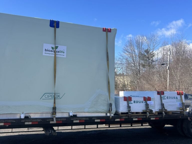bioaqualife: A flatbed truck transports large, strapped containers including a white item labeled "bioaqualife." Smaller boxes labeled "EDURA" sit beside it. Trees and a blue sky are visible in the background.
