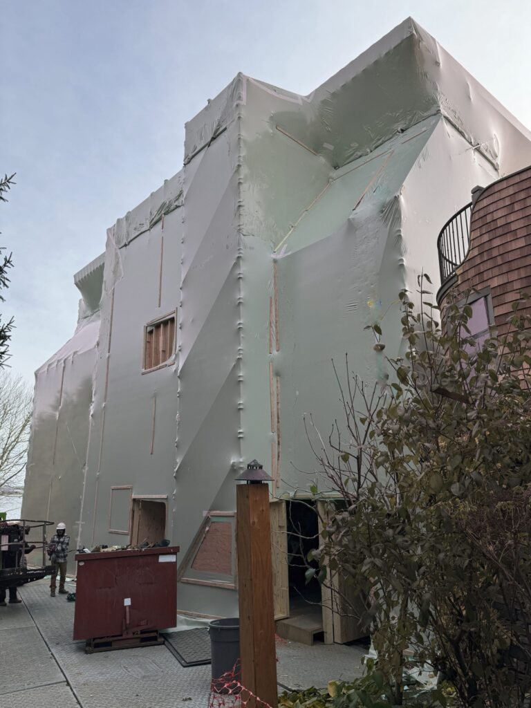 bioaqualife: A large building under construction is fully wrapped in pale plastic sheeting. The exterior is covered with seams and fastenings. Construction equipment and a red dumpster are on the sidewalk nearby.