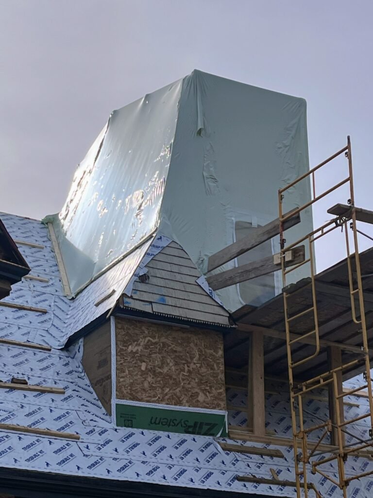bioaqualife: A rooftop under construction with scaffolding and a large chimney covered in plastic sheeting. The roof is partially covered with building wrap, and exposed plywood is visible on the walls.