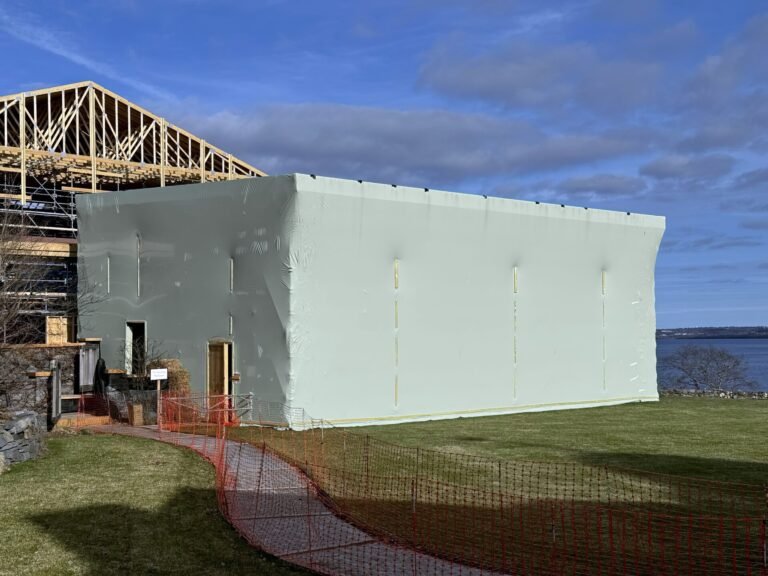 bioaqualife: A large building under construction is covered in white protective sheeting, with exposed framework visible on the roof. Orange safety fencing surrounds the area. There is a grassy lawn and water in the background.