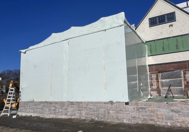 bioaqualife: A partially constructed house with white plastic sheets covering a section, stone foundation, ladders, and building materials visible outside; part of the house exterior is unfinished under a clear blue sky.