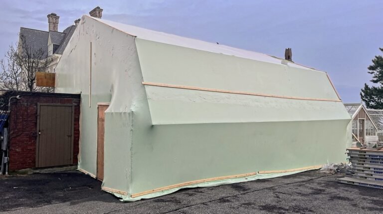 bioaqualife: A large structure covered in a pale, light green protective tarp is set up next to a brick building and a greenhouse. Wooden planks secure the tarp around the structure, which stands on an asphalt surface.