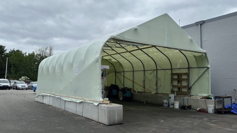 bioaqualife: A pale green, temporary shelter with a curved roof stands on a concrete base in a parking lot. Various tools and supplies are stored inside; cars and trees are visible in the background under a cloudy sky.