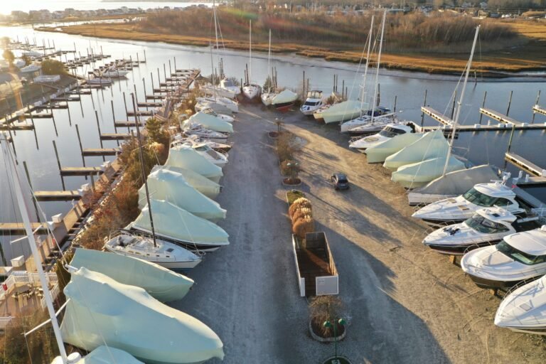 bioaqualife: Aerial view of a marina with boats covered in protective tarps for storage along both sides of a central gravel path, near docks and water, with buildings and trees in the background.