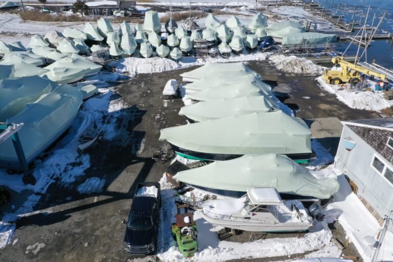 bioaqualife: Aerial view of boats covered in protective tarps and surrounded by snow at a marina in winter. Cars are parked nearby and the water is visible on the right side.