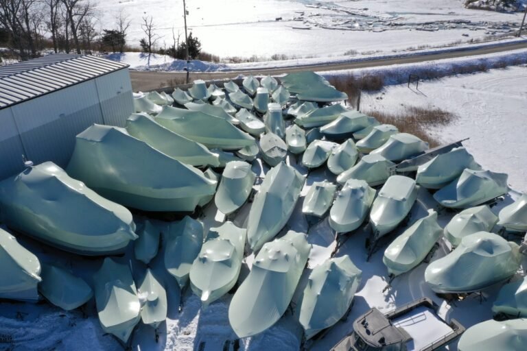 bioaqualife: Aerial view of many boats covered in shrink wrap for winter storage, arranged closely together on snow near a building and a road, with a frozen landscape in the background.