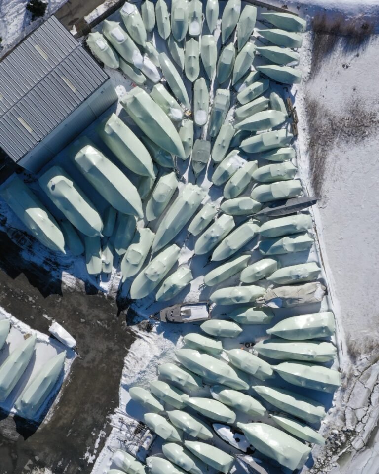 bioaqualife: Aerial view of many boats covered in white tarps, arranged closely together on a snow-covered ground near a building during winter.