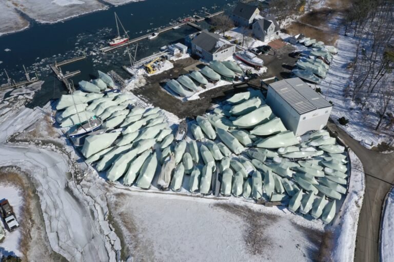 bioaqualife: Aerial view of a snowy boatyard with dozens of boats covered in white tarps, surrounded by ice and snow, near a partially frozen marina. Buildings and a few cars are visible nearby.