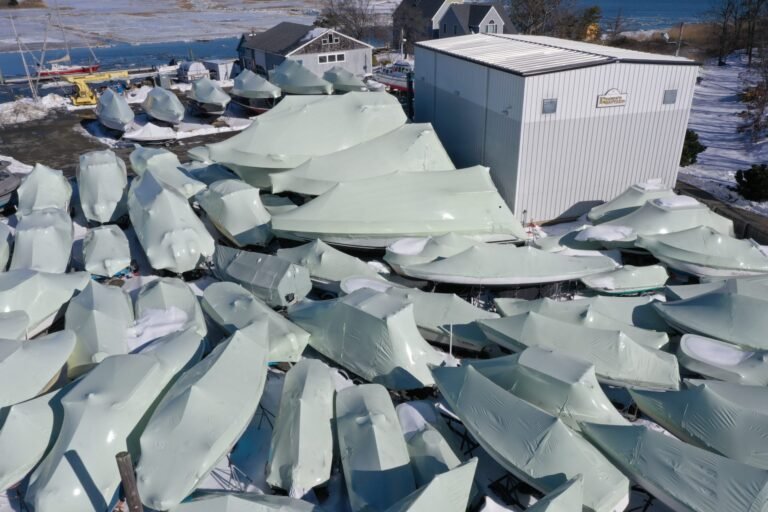 bioaqualife: Aerial view of numerous boats covered in white protective tarps, arranged closely together beside a white building on a snowy lot near a body of water.