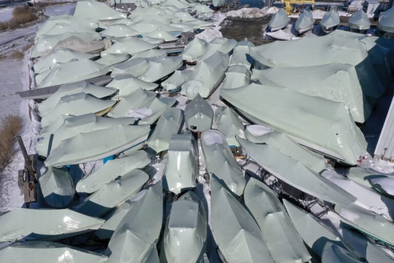 bioaqualife: Aerial view of numerous boats covered in white protective shrink wrap, arranged closely together in a snowy outdoor storage area during winter.