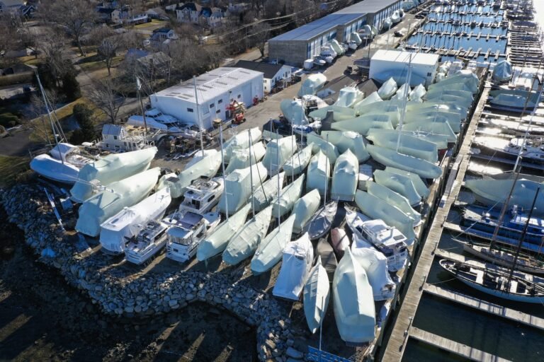 bioaqualife: Aerial view of a marina with numerous boats covered in white protective shrink wrap, stored on land next to a dock and a rocky shoreline; buildings and trees are visible in the background.